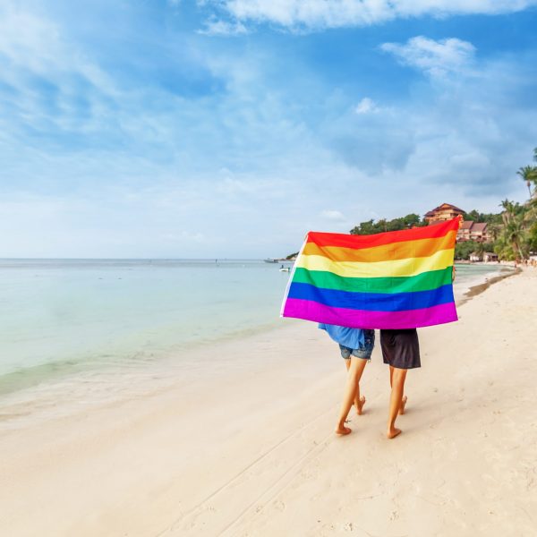 Young lesbian couple walks on a bright tropical beach with a rainbow flag, equal rights, symbol of lgbt community